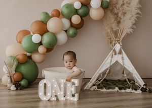 Neutral-toned balloon arch with sage green and brown decorations creates an elegant backdrop for a cake smash photo session.