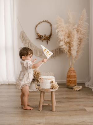 Adorable toddler in white outfit explores first birthday cake setup with pampas grass decor and woven stool.