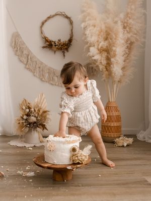 A small child in white lace romper reaches toward a rustic cake on wooden stand, with pampas grass and boho decor.