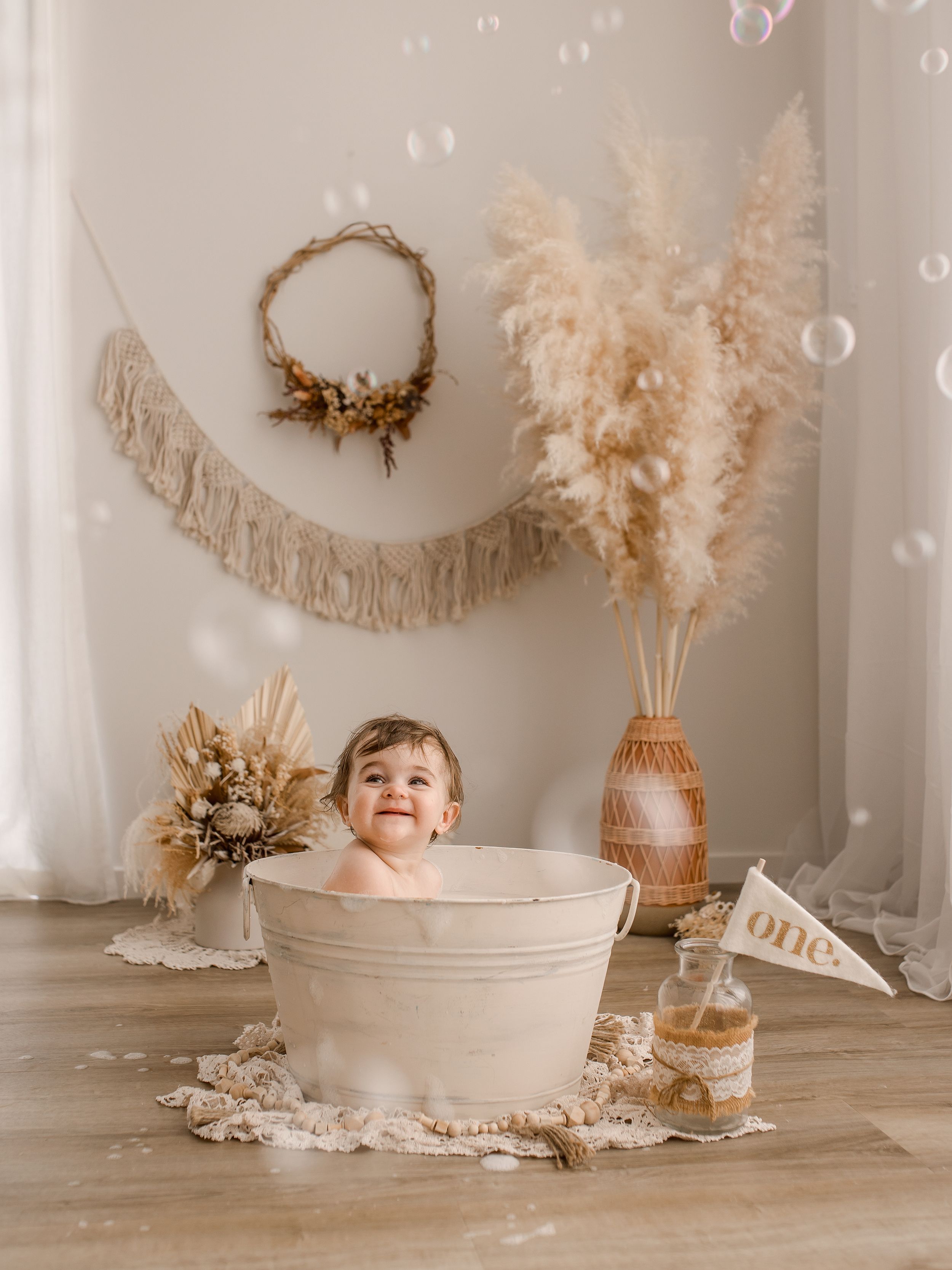 A rustic neutral-toned nursery setup with pampas grass, macrame wall decor, and child in white washtub for milestone photos.