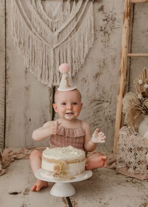 Baby in pink party outfit sits on floor with white cake during first birthday photo session against macrame backdrop.