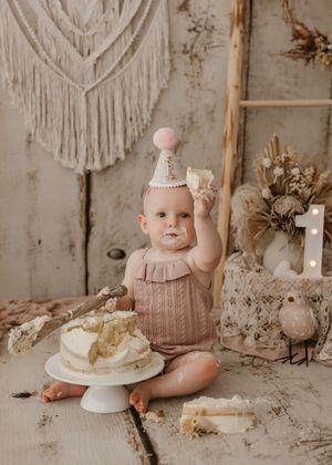 A baby in a pink dress sits on a rustic backdrop with vintage decor and a cake smash setup for first birthday photos.