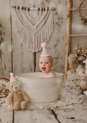 A happy baby sits in a white wash basin surrounded by rustic decor and bunny decorations in a neutral-toned setting.