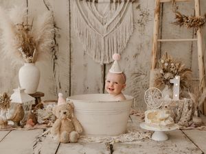 A rustic first birthday setup with vintage decor, pampas grass, teddy bear, and white bathtub in a neutral color scheme.
