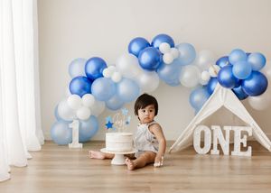 Baby's first birthday cake smash with blue balloon garland, white tipi, and ONE sign on hardwood floor.