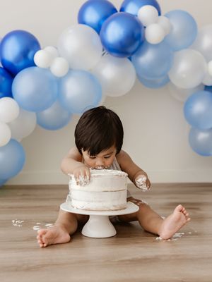 Baby boy smashing white birthday cake surrounded by blue and white balloon garland during first birthday cake smash photo session.