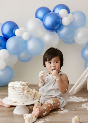 Baby's first birthday cake smash with blue and white balloon garland backdrop, messy frosting on hands and face.