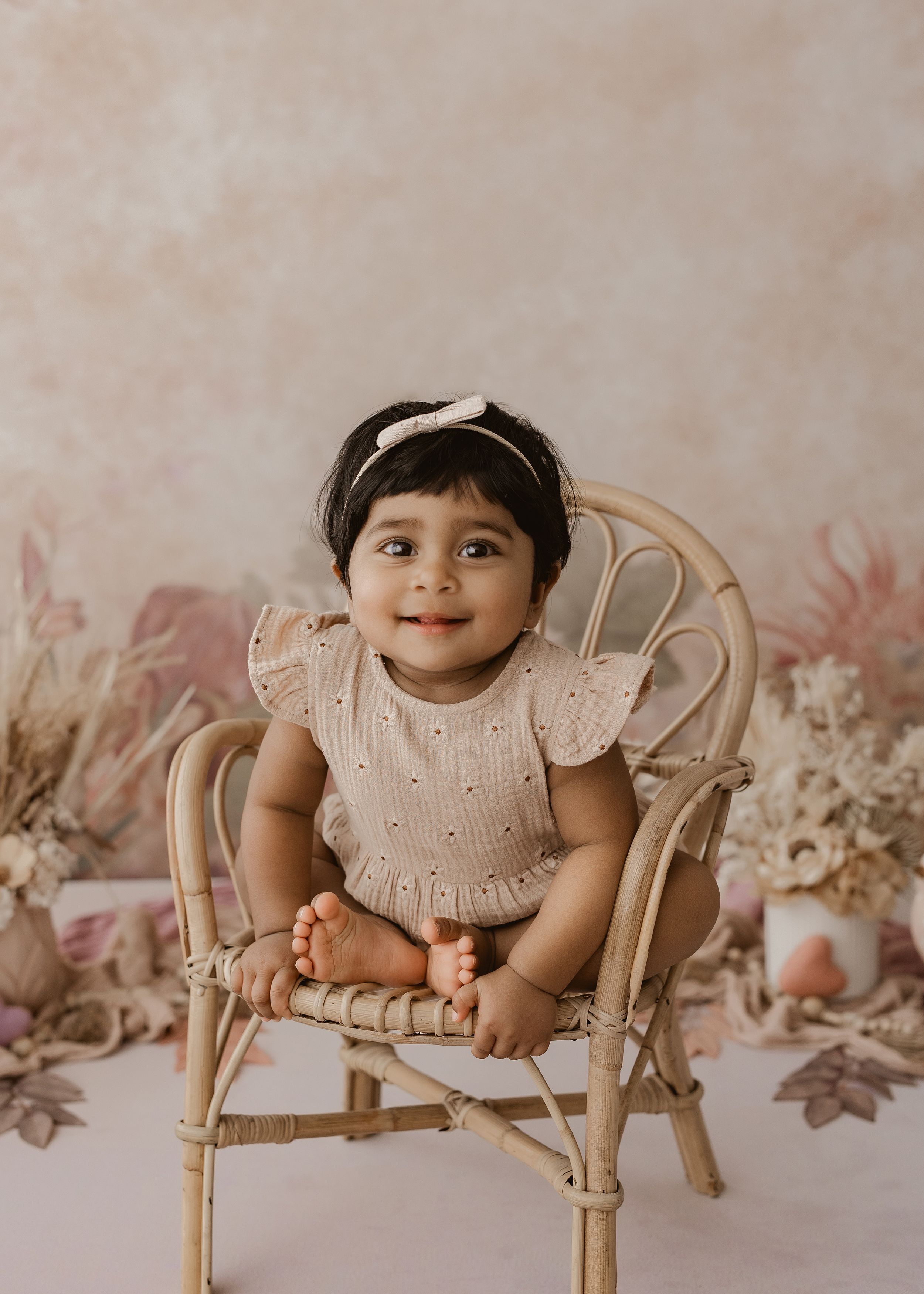 A baby in a white eyelet dress poses on a vintage wooden chair against a soft pink floral backdrop.