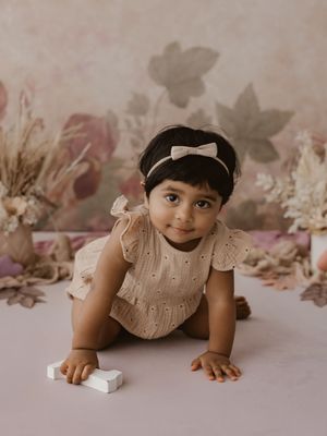 A small child in a white knit dress lies on a gray floor with a dreamy pink floral backdrop.