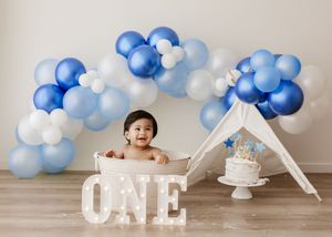 Baby's first birthday photoshoot with blue balloon arch, ONE marquee letters, teepee, and smash cake on white background.