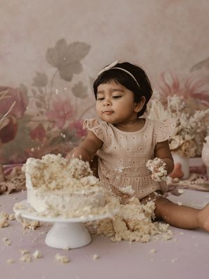 A cake smash photo session shows a small child in a cream dress making a delightful mess with white frosted cake.