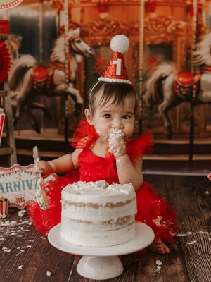 A red and white themed cake smash photo session featuring a classic white cake on a wooden surface with carnival carousel decor.