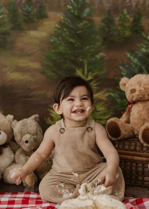 Smiling baby in tan overalls enjoys a cake smash surrounded by teddy bears and Christmas trees backdrop.