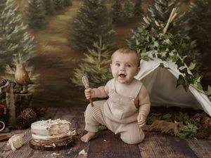 Baby in beige knit overalls smashing first birthday cake outdoors with woodland forest photography backdrop.