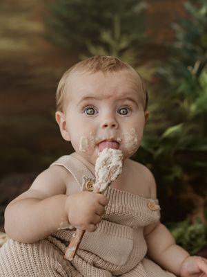 Adorable baby in beige knit romper eating cake with wide eyes, green foliage backdrop, first birthday smash cake photo.