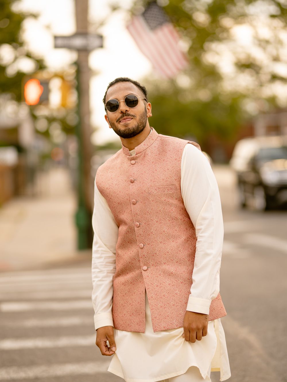 A man in formal white kurta and peach-colored vest poses outdoors on a city street at sunset.