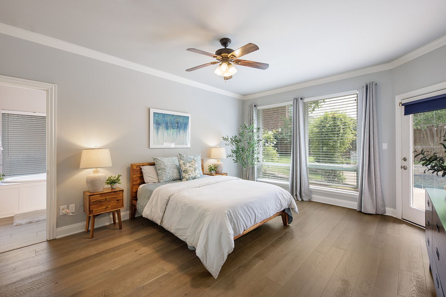 Modern bedroom with hardwood floors, white bedding, and large windows showcasing natural light and ceiling fan.