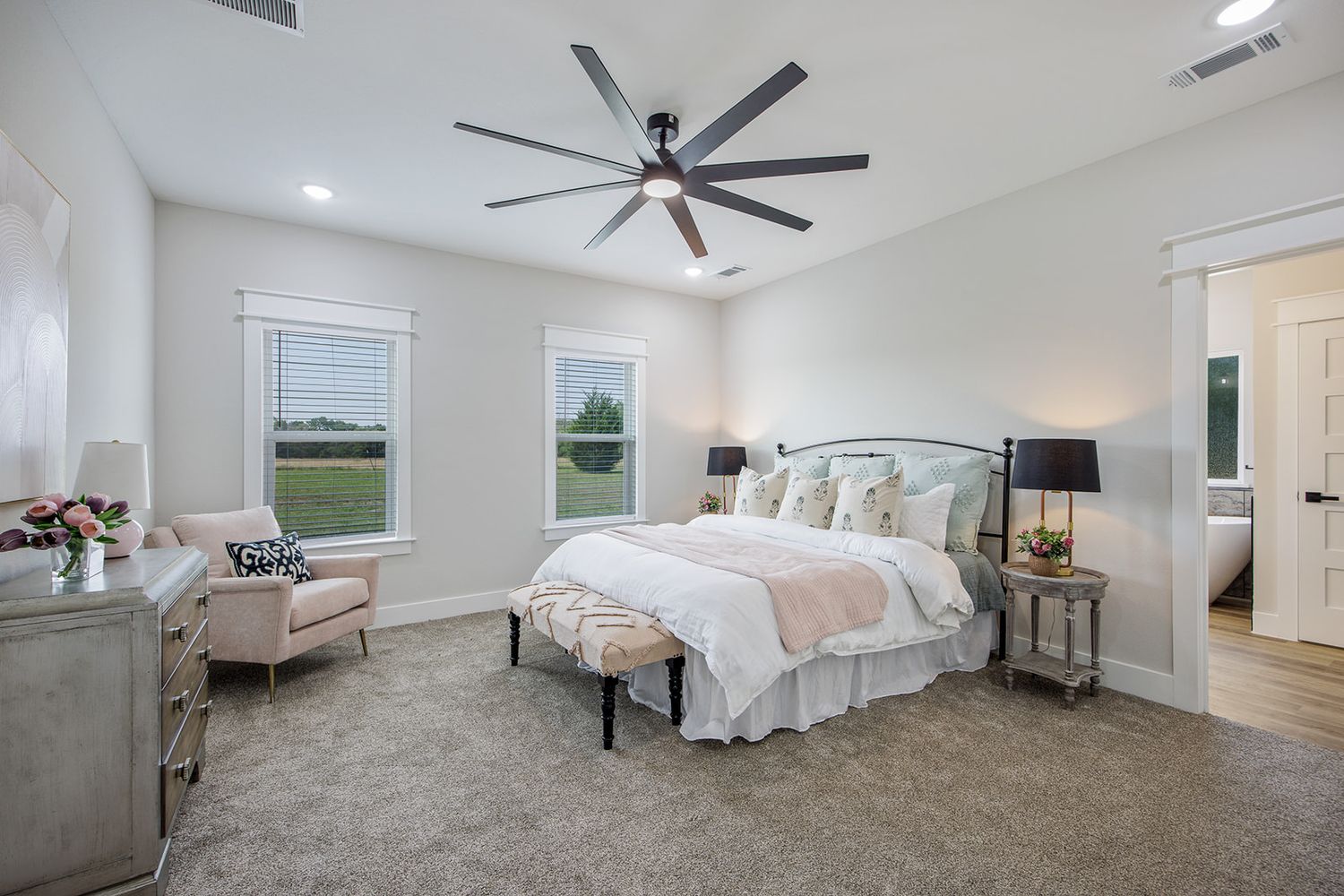 Bright master bedroom with luxurious white bedding, gray carpet, ceiling fan and large windows offering countryside views.