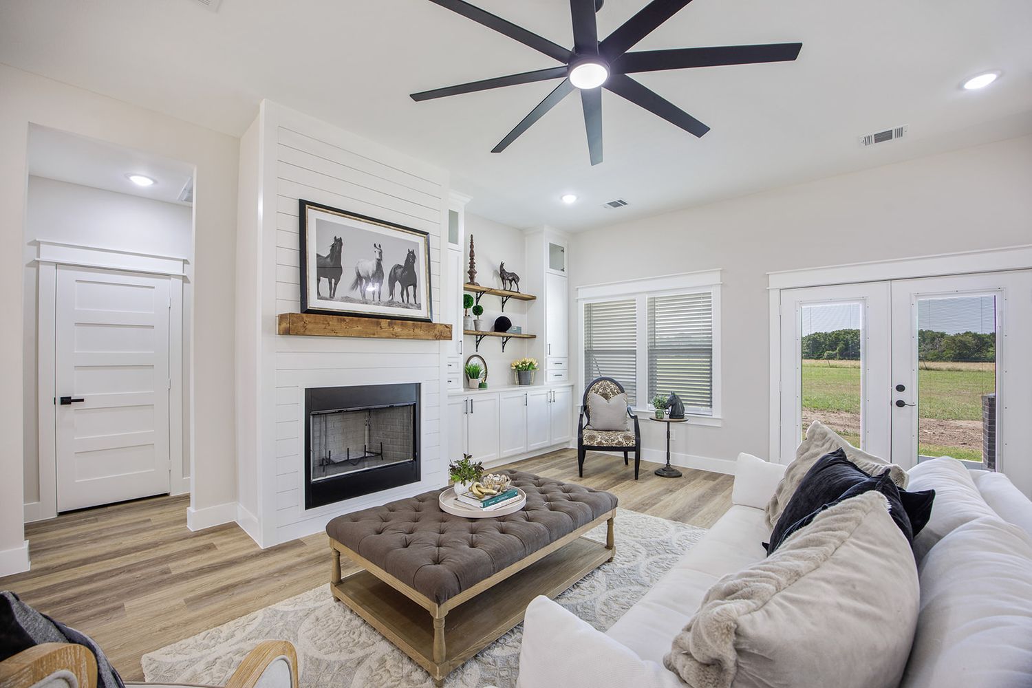 Modern farmhouse living room with white walls, floating shelves, wood mantle fireplace and large ceiling fan.