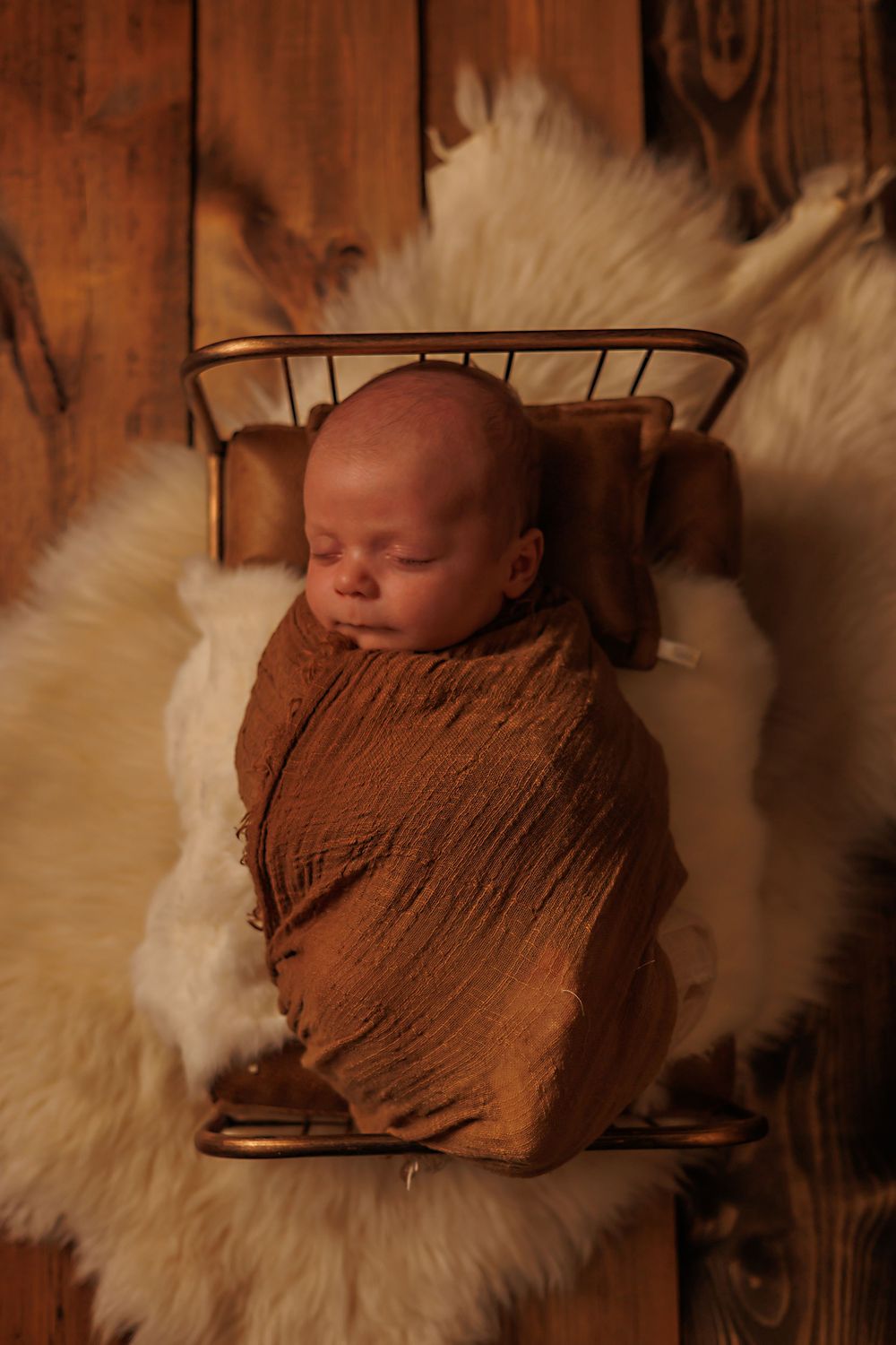 Sleeping newborn wrapped in brown fabric on white fur with rustic wood background.
