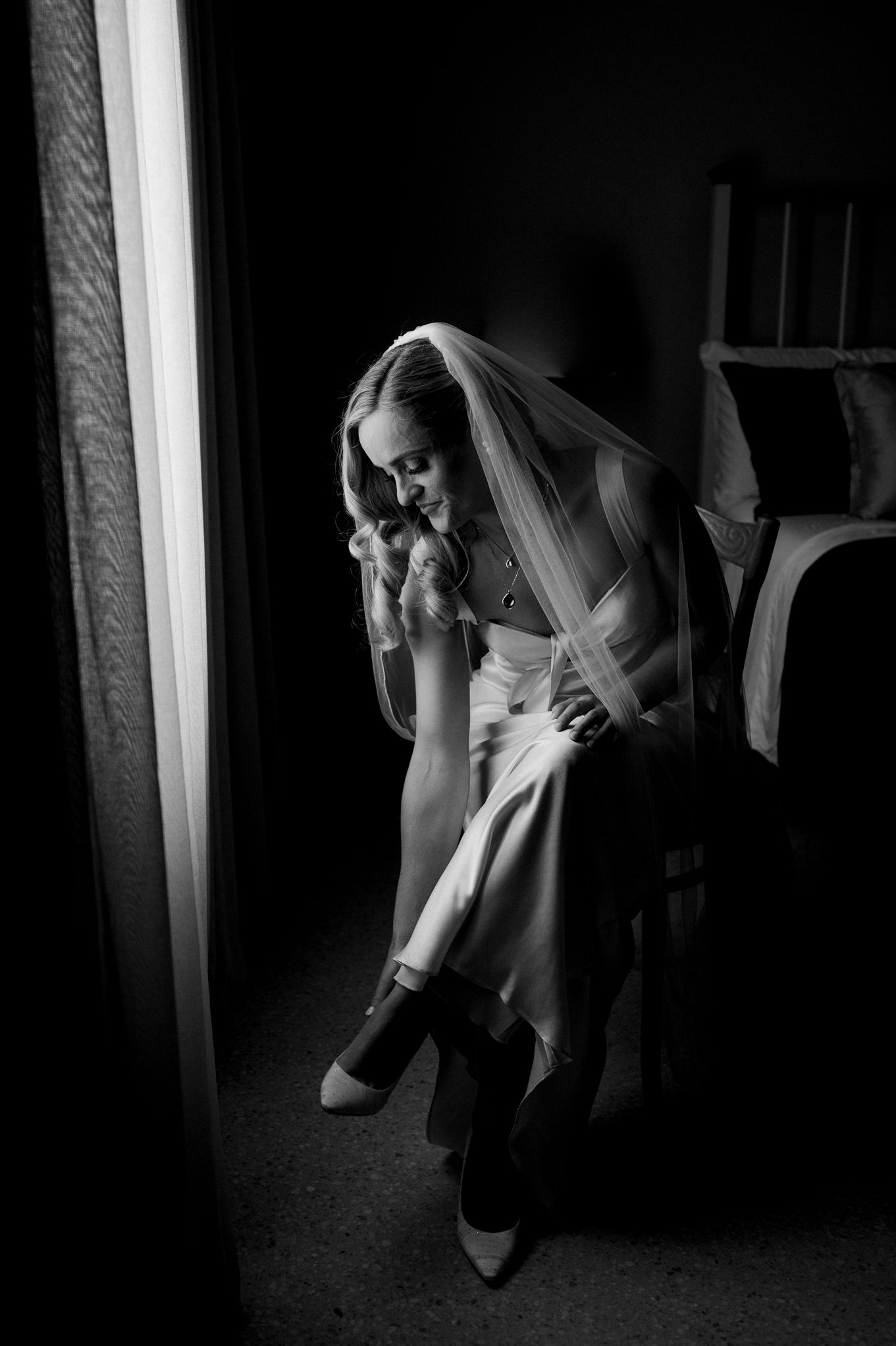 Dramatic black and white image of a bride adjusting her shoe while sitting in moody window light.