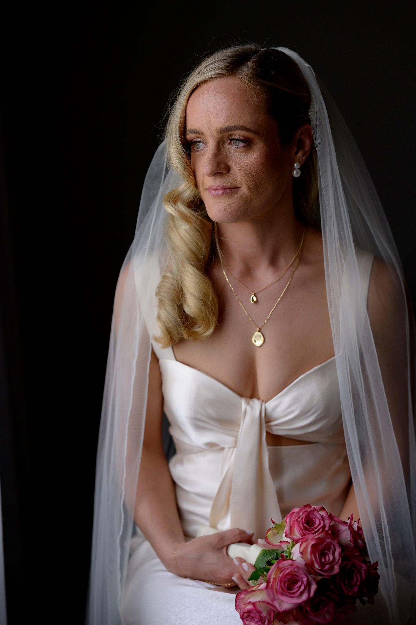 A bride in a white wedding dress holds a pink rose bouquet while wearing a long veil against a dark background.