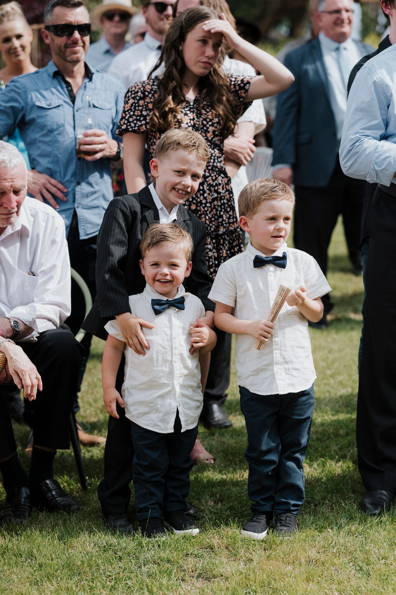 Young children laughing and playing on the grass during an outdoor wedding celebration with guests watching in the background.