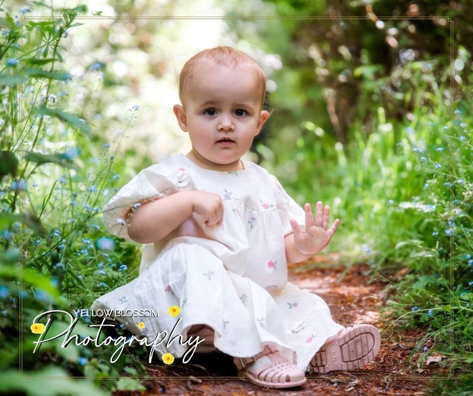 Small child wearing white dress sits on garden path surrounded by greenery in summer portrait setting.