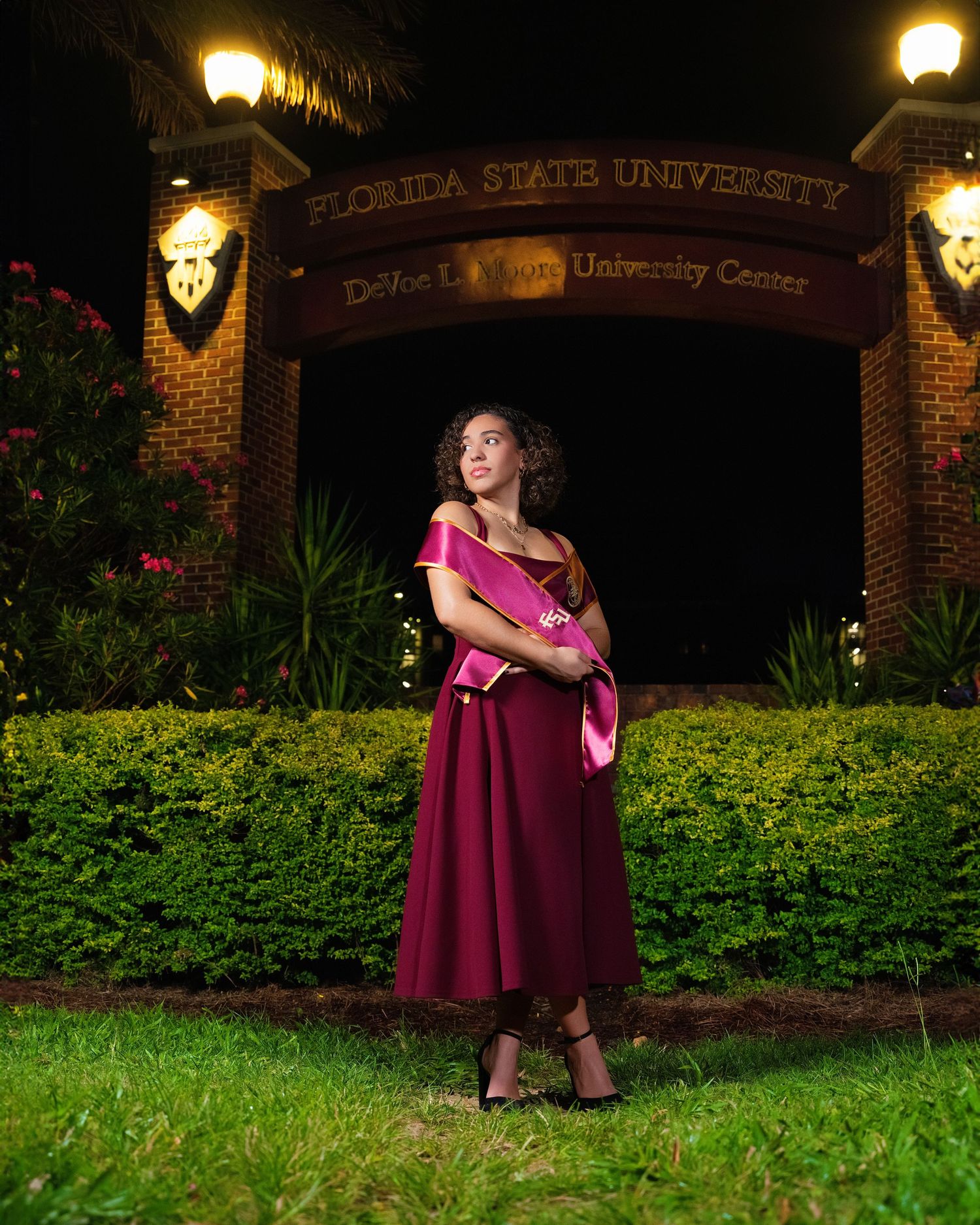 A person poses in a burgundy dress near an illuminated brick entryway on a summer evening in Florida.