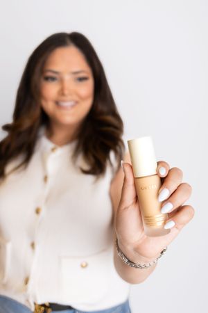 A hand holds a premium makeup foundation bottle against a white background while wearing a cream colored blouse.