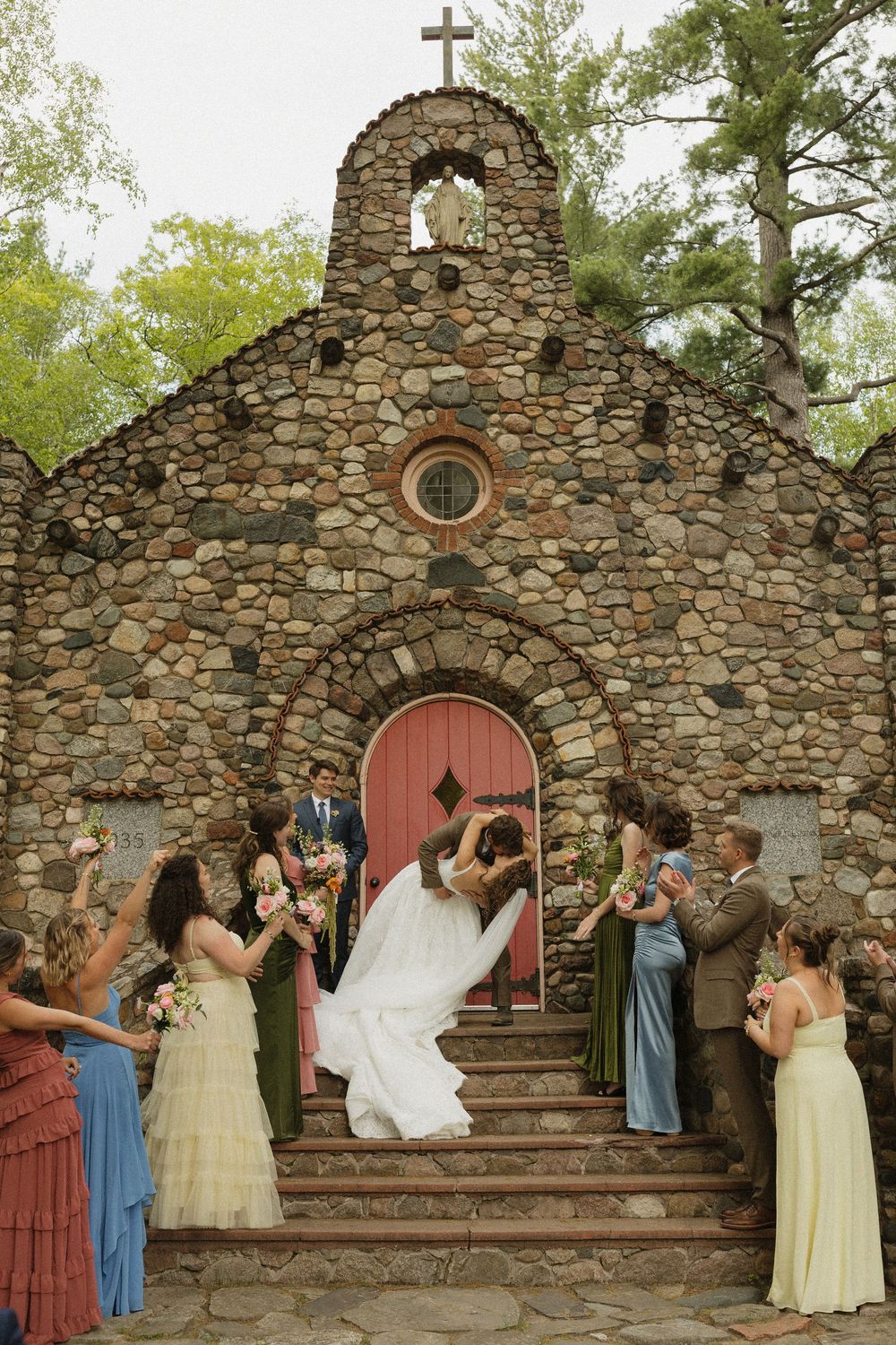 Bride and groom share a dip kiss on stone chapel steps surrounded by wedding party holding colorful bouquets.