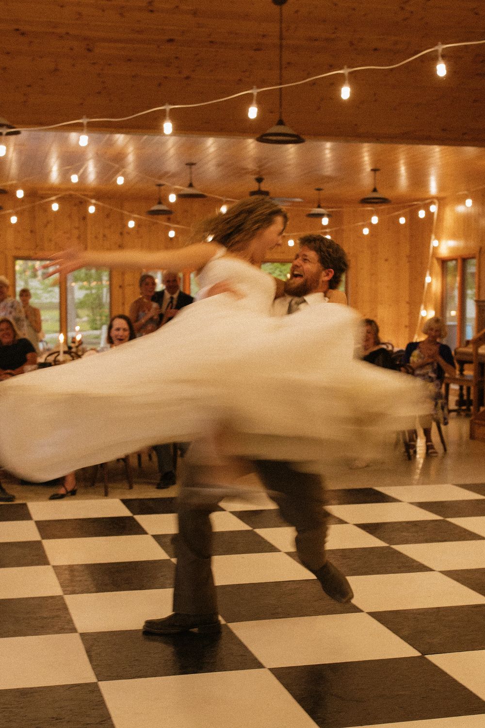 Bride and groom share first dance, spinning on checkered floor in warmly lit rustic barn venue with guests watching.
