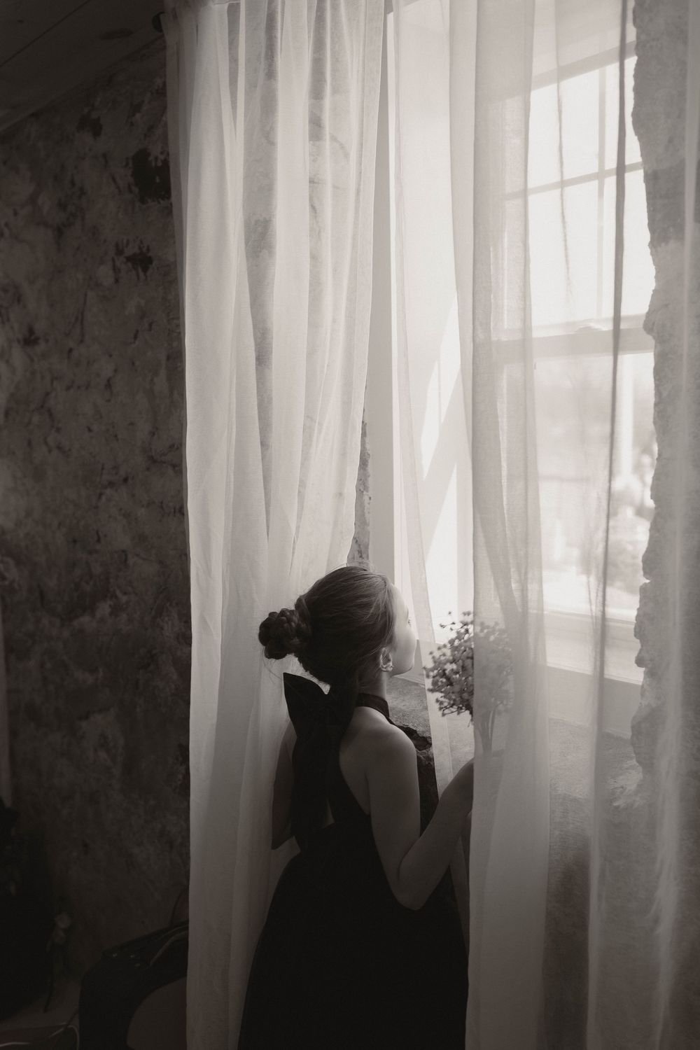 Silhouette through sheer curtains shows figure holding flowers in black and white photography.