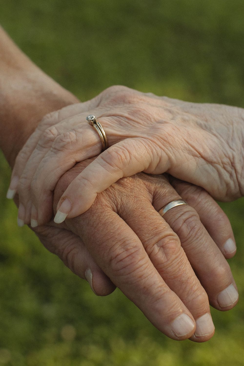 Close-up of weathered hands with wedding rings clasped together against a green background.