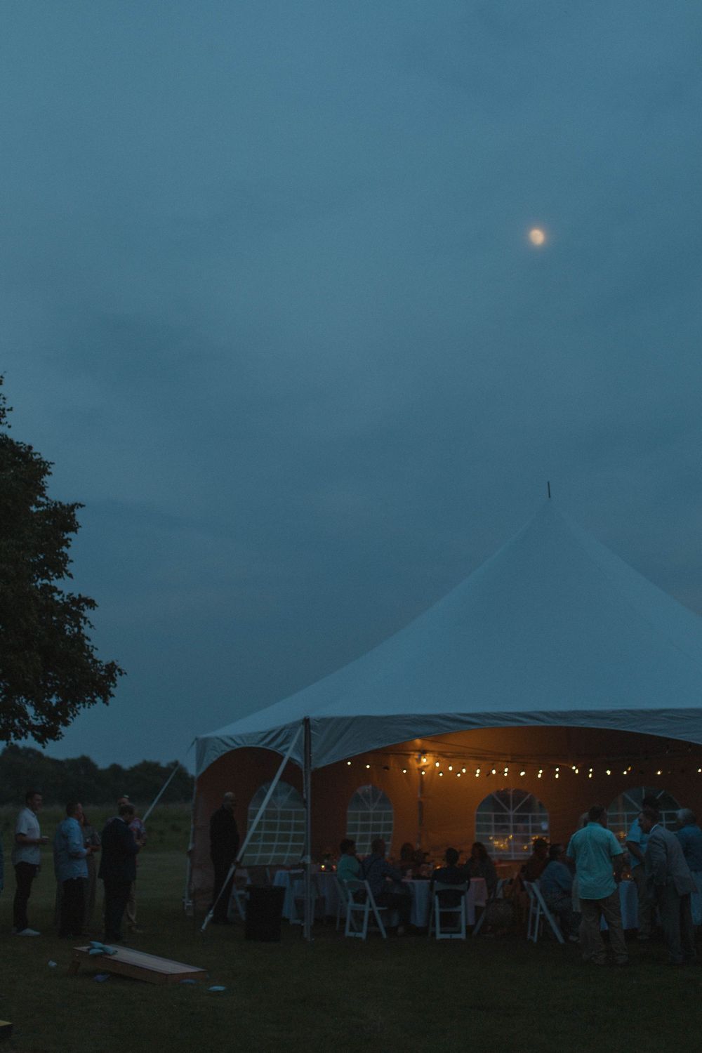 An illuminated wedding tent glows at dusk with string lights and guests mingling outside.