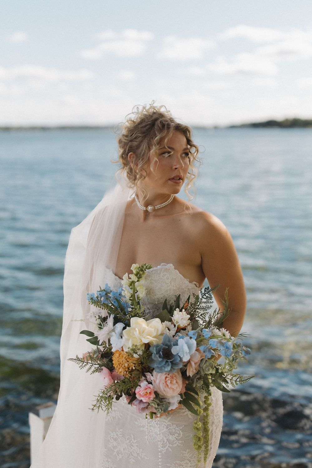 Bride in strapless gown holding colorful bouquet poses by waterfront with veil flowing in breeze.