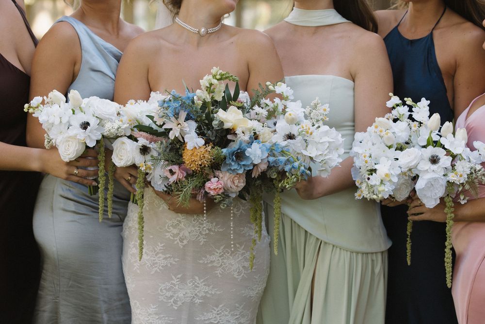 Row of bridesmaids holding pastel and white floral bouquets at outdoor wedding.