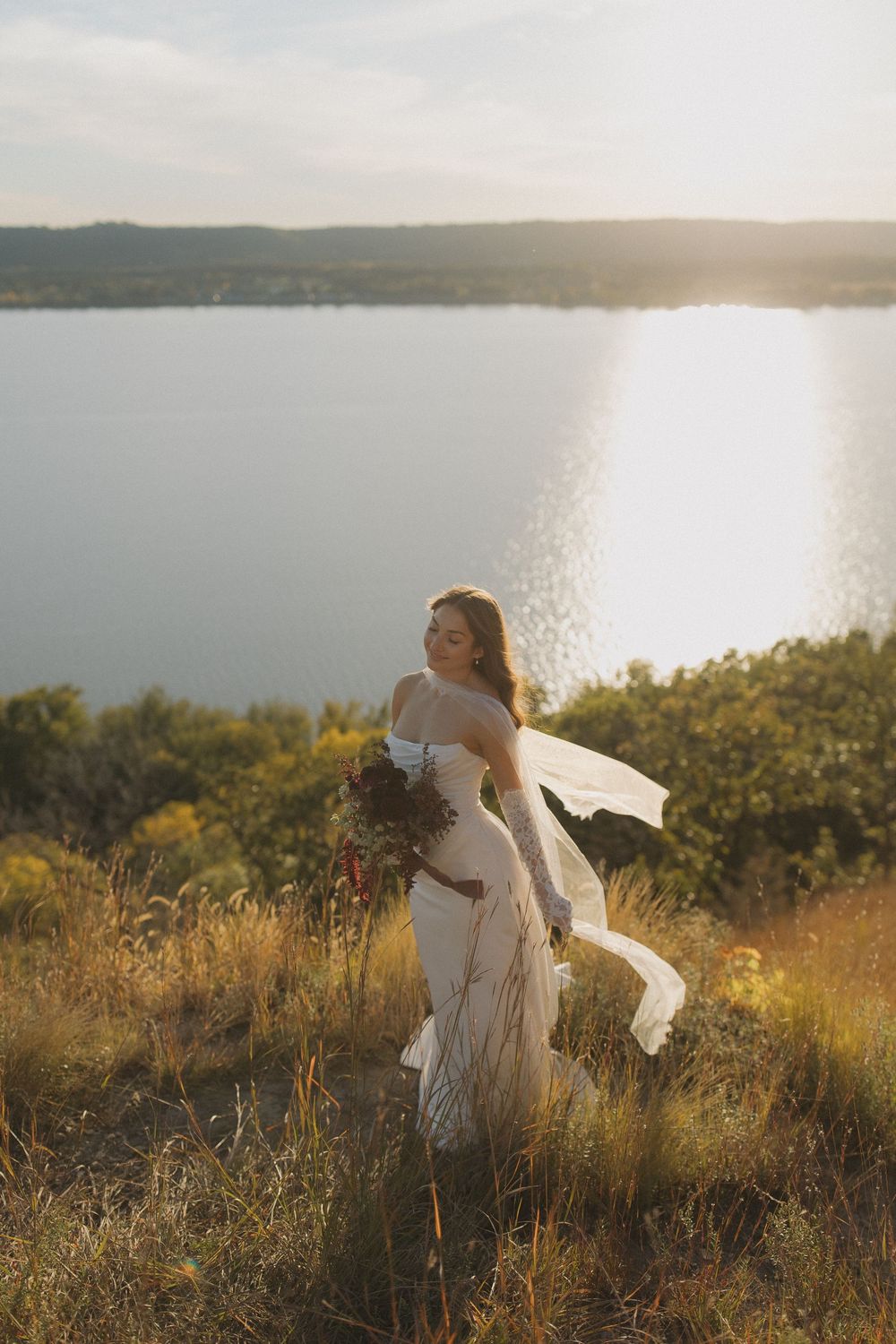 Person in white dress twirling on hilltop overlooking lake at sunset.