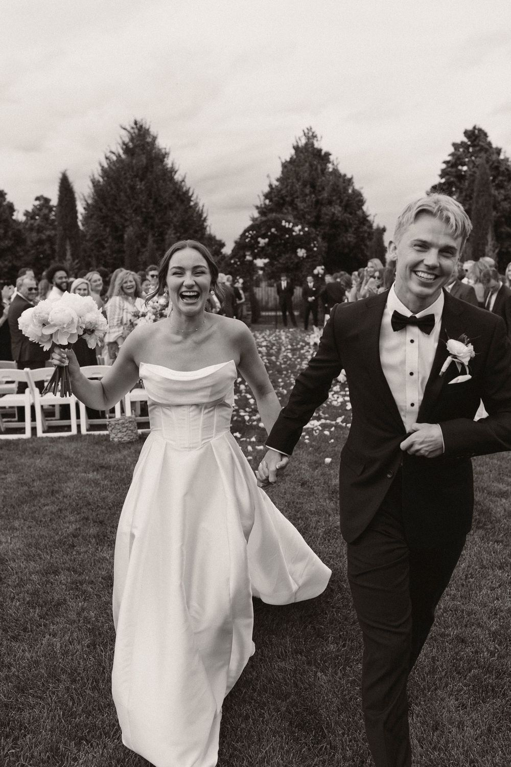 A joyful wedding couple celebrates while walking outdoors with white chairs visible in the background.