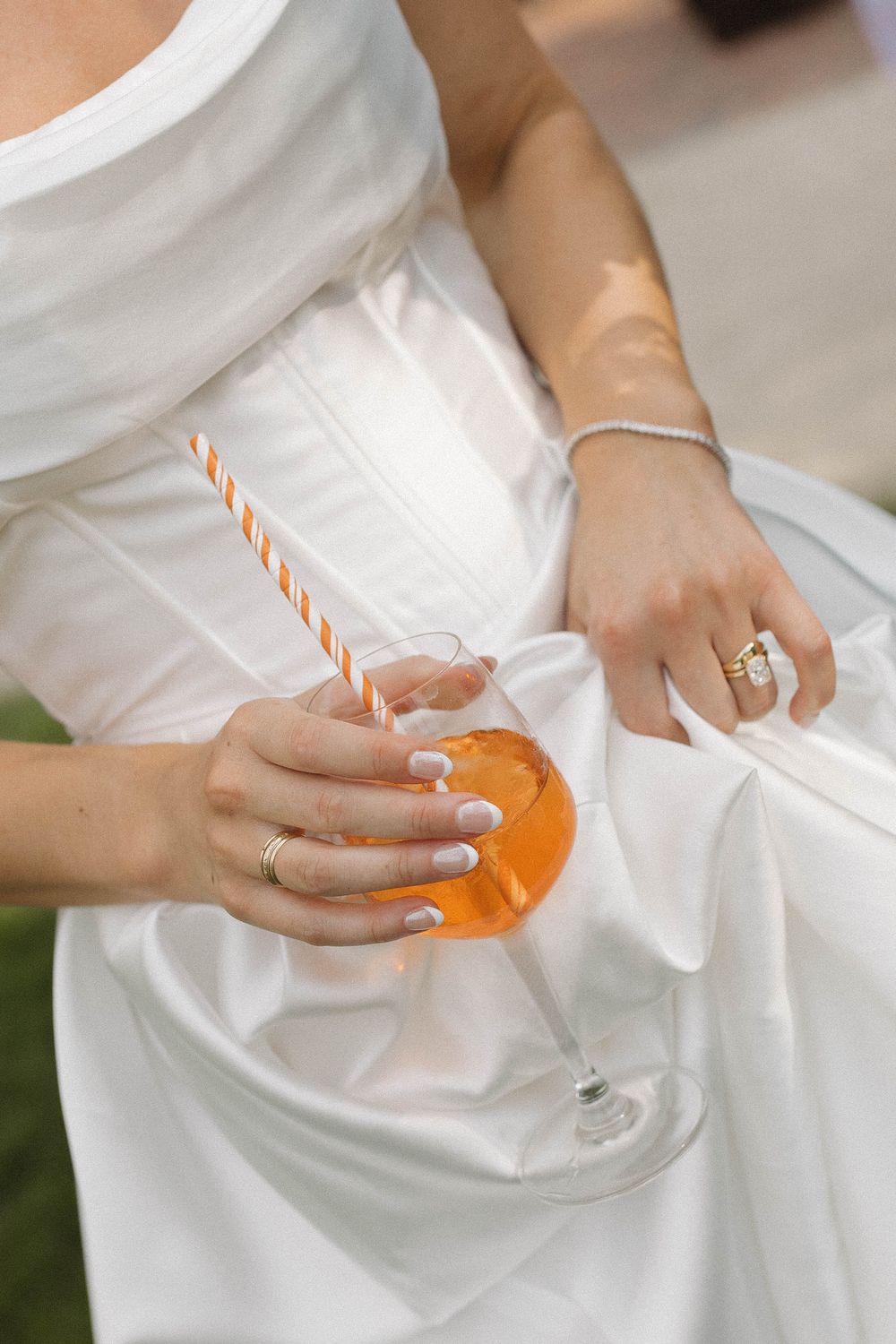 Close-up of hands holding an orange cocktail, showing wedding rings and white dress details.