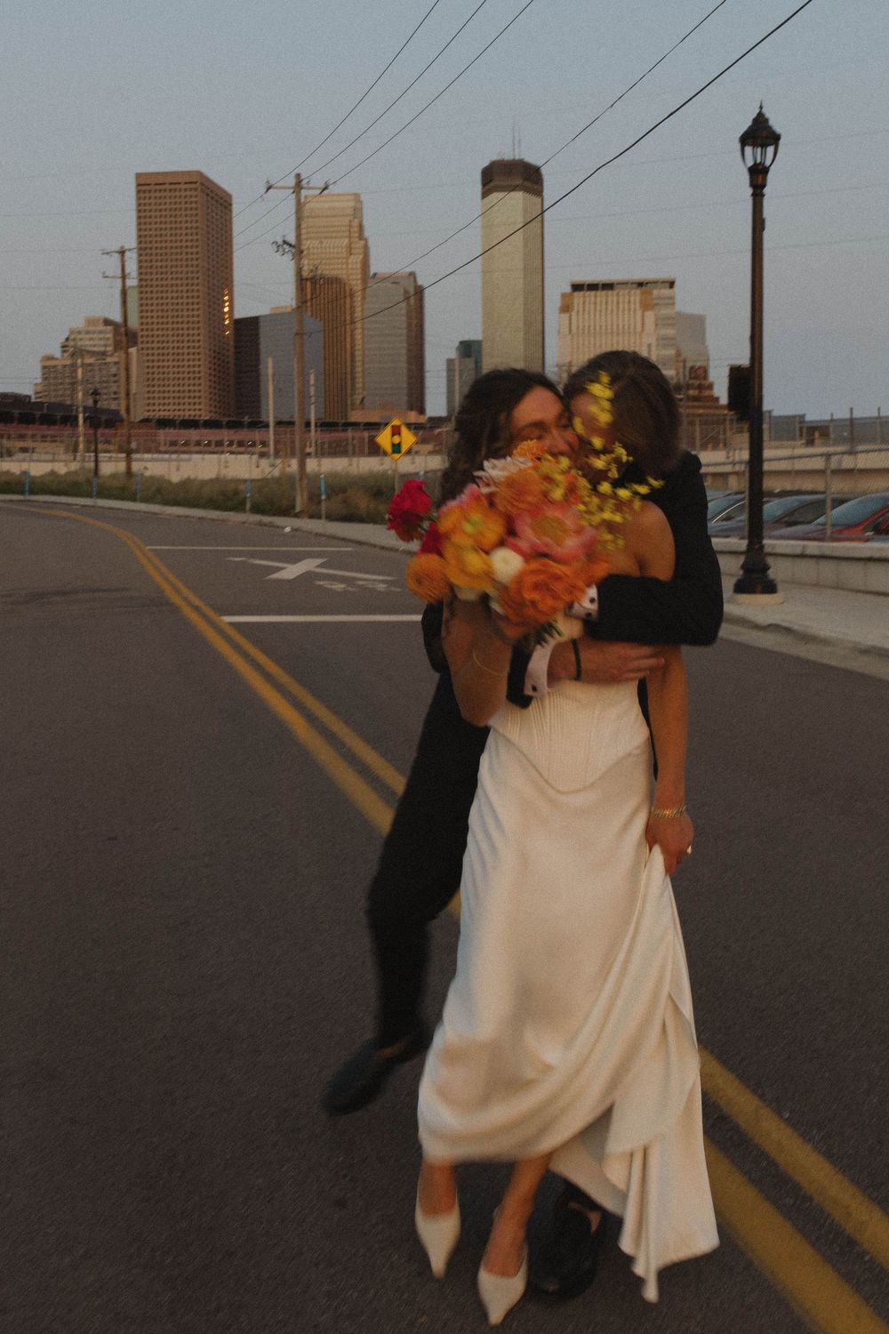 Bride in white gown holding orange bouquet embraced by groom on city street with skyline backdrop at dusk.