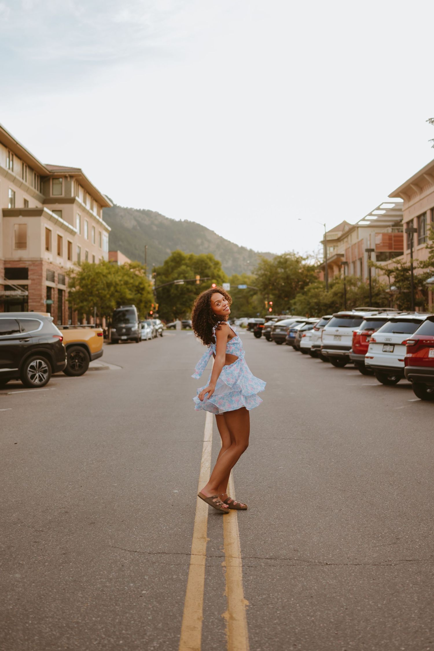 Carefree dancer in blue dress twirls on yellow line in middle of city street at sunset with mountains in background.