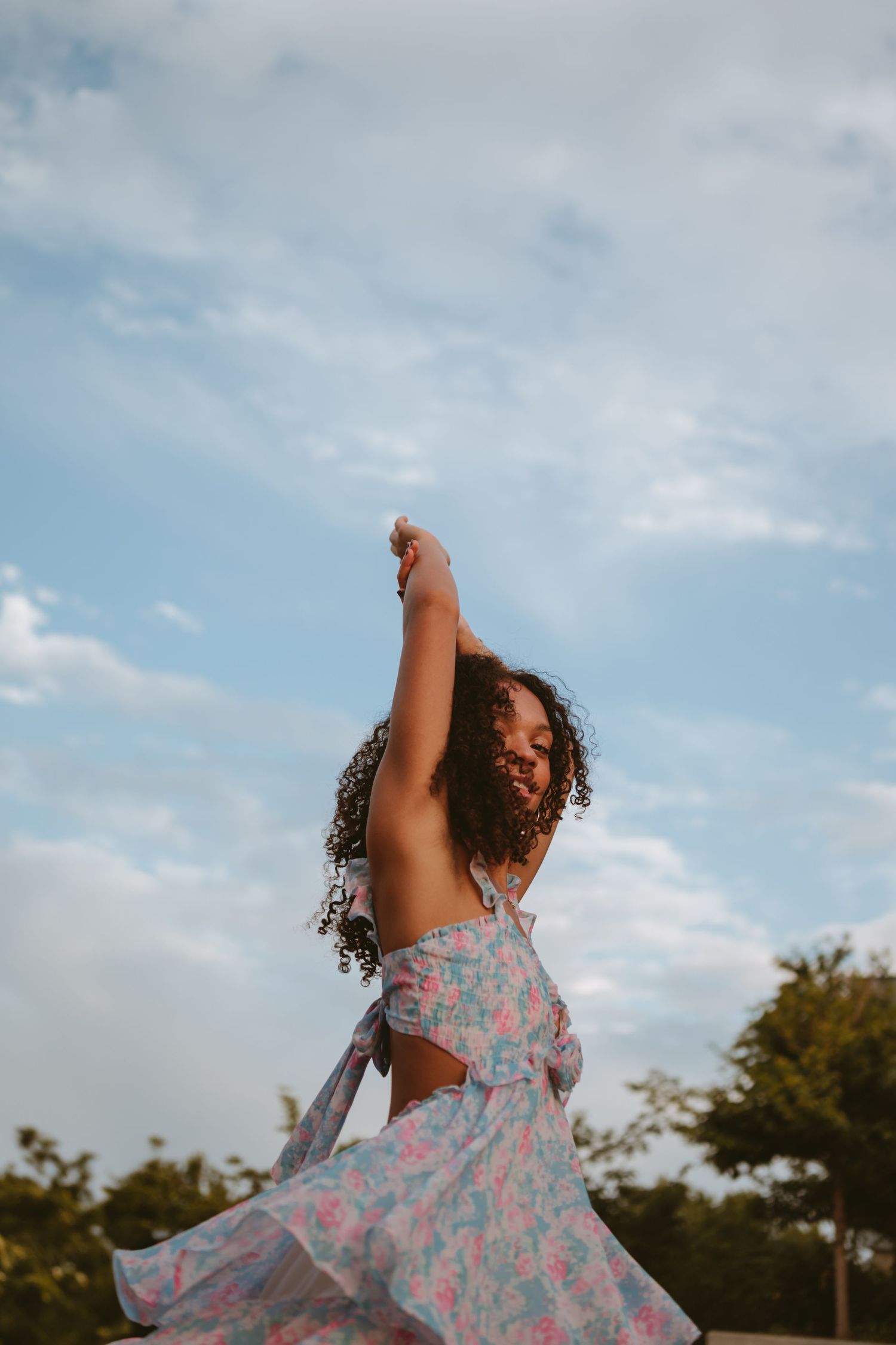 A person in a floral dress twirls joyfully outdoors against a blue sky with wispy clouds.