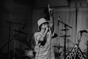 A performer in a striped shirt and baseball cap sings into a microphone on stage during a concert performance.