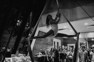 An aerial performer gracefully suspends from silk fabric during an indoor performance with audience members watching.