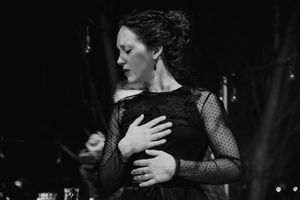 A dramatic black and white performance shot of a performer in a lace long sleeve dress against a dark background.
