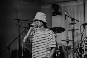 A performer in a striped shirt and bucket hat sings into a microphone on stage.