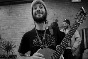 Musician holding acoustic guitar smiles while performing in front of brick wall.