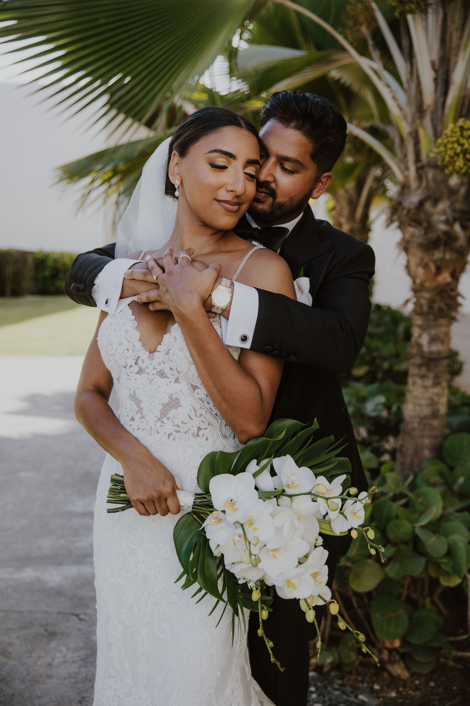 Elegant wedding couple embracing under palm trees with white orchid bouquet.