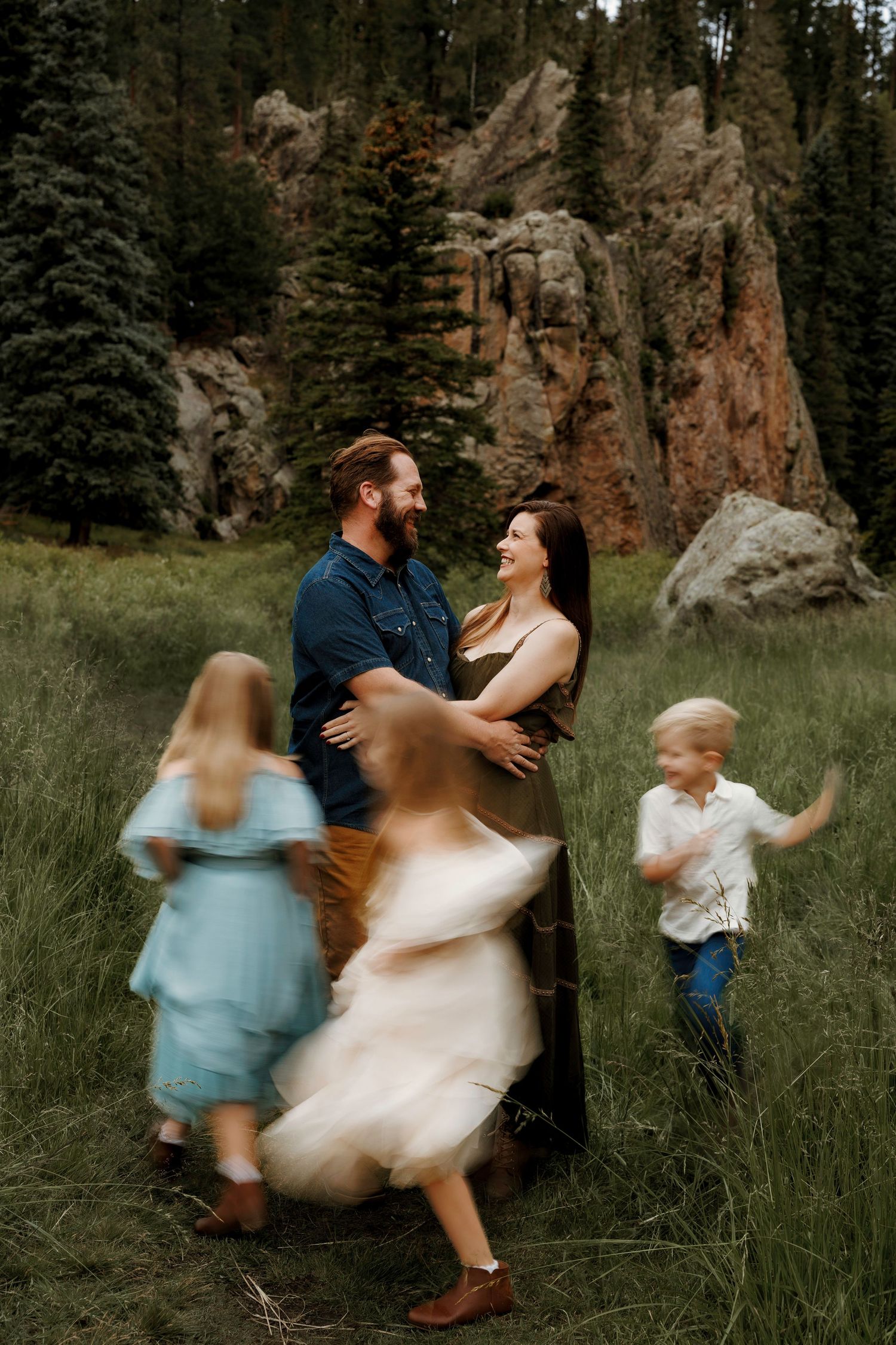Family with children playing in a mountain meadow surrounded by pine trees and rocky cliffs.
